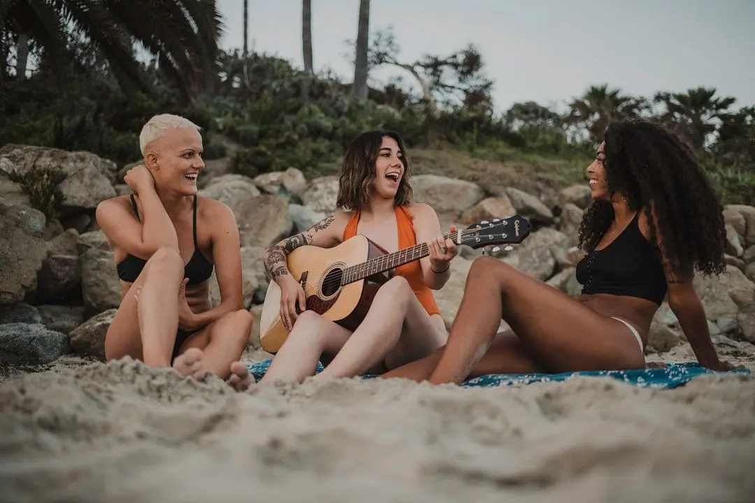 Three women sitting on the beach playing a guitar